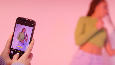 Studio-Shot-Of-Woman-Taking-Photo-Of-Friend-Dancing-On-Mobile-Phone-Against-Pink-Background-3